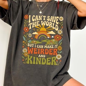 Pode incluir: Uma t-shirt de manga curta cinzento-carvão com uma impressão gráfica colorida que mostra um arco-íris, um sol e flores. O texto diz: I CAN'T SAVE THE WORLD BUT I CAN MAKE IT WEIRDER AND KINDER. O design inclui tons de laranja, amarelo, verde e vermelho.