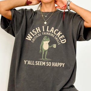 May include: A charcoal gray short-sleeve t-shirt featuring a graphic of a green frog holding a mug. The text reads WISH I LACKED CRITICAL THINKING SKILLS Y'ALL SEEM SO HAPPY in cream-colored lettering arched above and below the frog illustration.