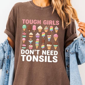 May include: A dark brown short-sleeve t-shirt featuring a graphic print of various colourful ice cream treats and the text "TOUGH GIRLS DON'T NEED TONSILS" in pink and white lettering. The shirt is worn with a light blue denim jacket.