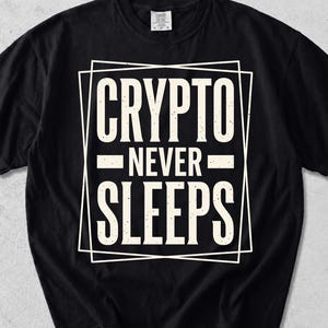 Op de afbeelding: Een zwart t-shirt met korte mouwen en een wit grafisch ontwerp met de tekst "CRYPTO NEVER SLEEPS" in een dubbel rechthoekig kader. De letters hebben een verweerde, getextureerde uitstraling en zijn gecentreerd op de borst geplaatst.