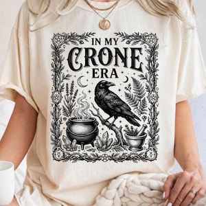 May include: Ivory-colored t-shirt with a black graphic design. The design features a raven perched on a branch, a cauldron, and the text "IN MY CRONE ERA" within a decorative frame. The shirt is made by Comfort Colors.