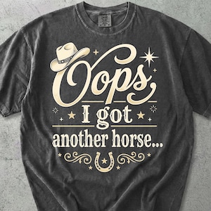May include: A charcoal gray short-sleeve t-shirt featuring a cream-colored graphic print. The design includes a cowboy hat icon, a horseshoe, and the text "Oops I got another horse..." in a decorative script and serif font with star accents.