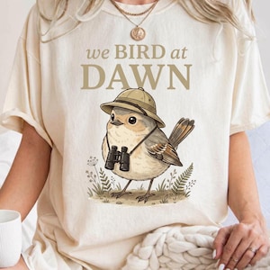 Könnte beinhalten: Ein elfenbeinfarbenes Kurzarm-T-Shirt mit einer Grafik eines kleinen Vogels, der einen Safarihut und ein Fernglas trägt. Der Text über dem Vogel lautet we BIRD at DAWN in beiger Schrift. Das T-Shirt hat einen Rundhalsausschnitt und eine lockere Passform.