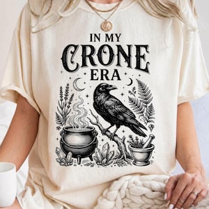 May include: An ivory short-sleeve t-shirt featuring a black graphic print with the text "IN MY CRONE ERA" above an illustration of a raven perched on a branch, a steaming cauldron, crystals, and botanical elements like ferns and lavender.