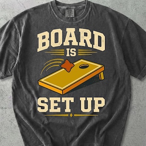 May include: A charcoal grey short-sleeve t-shirt featuring a graphic of a yellow cornhole board and a bean bag in flight. The text reads BOARD IS SET UP in a bold, distressed cream-coloured font, centred on the chest of the garment.