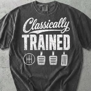 Może przedstawiać: Szara koszulka z napisem "Classically Trained" w kolorze białym, z grafiką zmiany biegów i pedałów. Koszulka ma okrągły dekolt i krótkie rękawy.