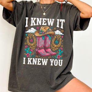 May include: A charcoal gray short-sleeve t-shirt featuring a graphic print of a cowboy hat, pink western boots, horseshoes, and cacti. The text reads I KNEW IT I KNEW YOU in white block letters. The design includes colorful floral accents.