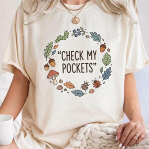 Peut inclure: T-shirt ivoire avec l'inscription "CHECK MY POCKETS" entourée d'une couronne de feuilles, de glands, d'un champignon et d'insectes. Le t-shirt est de la marque Comfort Colors.