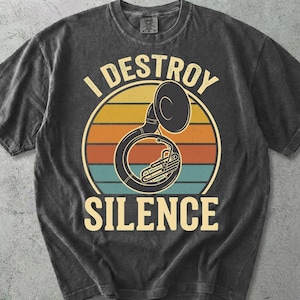 Op de afbeelding: Een antracietgrijs t-shirt met korte mouwen en een afbeelding van een tuba in het midden van een cirkel in zonsondergangkleuren met horizontale strepen. De tekst "I DESTROY SILENCE" staat in een vet, lichtgekleurd lettertype boven en onder de tuba.