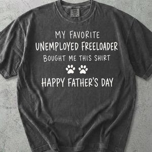 May include: Pepper-colored t-shirt with white text: "MY FAVORITE UNEMPLOYED FREELOADER BOUGHT ME THIS SHIRT" and "HAPPY FATHER'S DAY" with paw prints. The shirt has a crew neck and short sleeves.