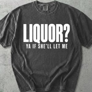 May include: A dark gray t-shirt with the text "LIQUOR? YA IF SHE'LL LET ME" in white block letters. The shirt has a crew neck and short sleeves. The fabric appears to be pre-washed for a vintage look.