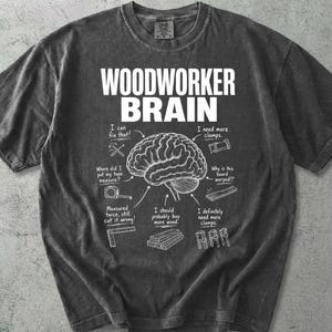 May include: Charcoal grey t-shirt with the text "WOODWORKER BRAIN" in white. The shirt displays a brain diagram with woodworking-related phrases and illustrations, including tools and wood pieces.