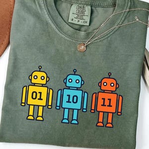 May include: Moss green t-shirt with a graphic of three robots in yellow, blue, and orange. Each robot has a number: 01, 10, and 11. The shirt has a crew neck and short sleeves. A gold necklace is draped across the shirt.