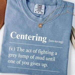Könnte beinhalten: Ein blaues Jeans-T-Shirt mit dem Wort "Centering" in Weiß und einer Definition. Eine goldene Halskette mit Anhänger liegt auf dem Shirt. Das Shirt hat ein Comfort Colors-Etikett.