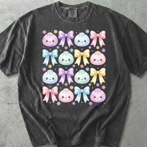 May include: A charcoal gray short-sleeve t-shirt featuring a repeating pattern of pastel-colored cartoon dumplings with smiling faces and colorful bows. The design includes small flower accents interspersed between the dumplings and bows.
