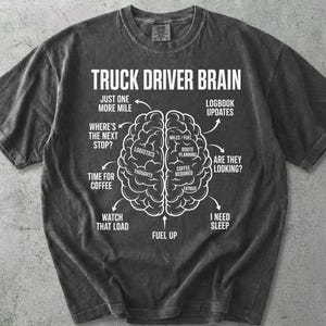 May include: Dark gray t-shirt with the text "TRUCK DRIVER BRAIN" and a brain diagram. The diagram includes phrases like "Logistics," "Fuel Up," and "I Need Sleep." The shirt is a casual wear item.