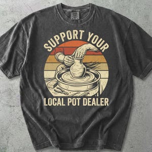 Puede incluir: Una camiseta gris carbón de manga corta con un gráfico circular sobre un fondo de atardecer. El diseño muestra manos dando forma a una vasija de barro en un torno de alfarero, con el texto "SUPPORT YOUR LOCAL POT DEALER" en letra negrita.