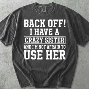 Puede incluir: Una camiseta de manga corta en color gris carbón con letras blancas en bloque que dicen: BACK OFF! I HAVE A CRAZY SISTER AND I'M NOT AFRAID TO USE HER. La prenda tiene un cuello redondo y una textura lavada de estilo vintage.