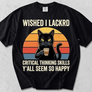 Könnte beinhalten: Ein schwarzes Kurzarm-T-Shirt mit einer Grafik eines schwarzen Katers, der eine Kaffeetasse vor einem Sonnenuntergangskreis hält. Der Text lautet: WISHED I LACKRD CRITICAL THINKING SKILLS Y'ALL SEEM SO HAPPY. Das Design verwendet gelbe, orange und blaue Töne.