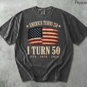 May include: A dark gray t-shirt with a distressed American flag graphic. The text reads "AMERICA TURNS 250" above the flag and "I TURN 50" below, with the years 1776-1976-2026. The shirt has a crew neck and short sleeves.