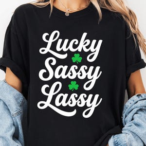 May include: Black t-shirt with the text "Lucky Sassy Lassy" in white script, accented with green shamrocks. The shirt is a casual style, suitable for St. Patrick's Day or everyday wear.
