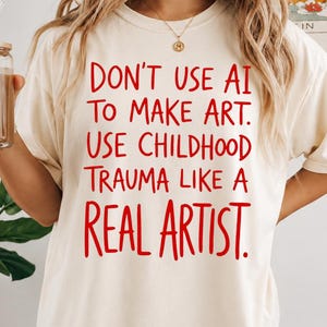 Può includere: Una t-shirt color avorio a maniche corte con testo rosso che recita: DON'T USE AI TO MAKE ART. USE CHILDHOOD TRAUMA LIKE A REAL ARTIST. La maglia presenta un girocollo e una vestibilità ampia, con un carattere in stile scritto a mano in rosso.