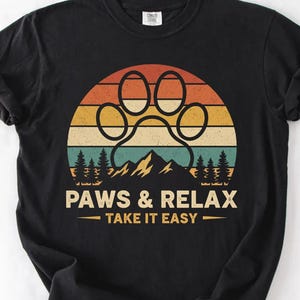 以下が含まれることがあります： 黒いTシャツ。夕焼けのデザインの上に足跡のグラフィックがあり、山と木が描かれています。テキストは「PAWS & RELAX - TAKE IT EASY」と書かれています。