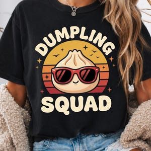 May include: Black t-shirt with a graphic of a dumpling wearing sunglasses, the text "DUMPLING SQUAD" in a circular design, and the words "Comfort Colors" in the upper left corner. The design features a sunset colour scheme.