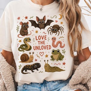 Op de afbeelding: Ivoor kleurig T-shirt met de tekst "Love the Unloved" en illustraties van een vleermuis, spin, slang, slak, kikker en andere wezens. De woorden "Comfort Colors" staan bovenaan het shirt.