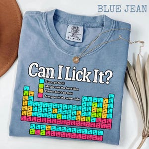 Peut inclure: Un t-shirt bleu jean avec un graphique de tableau périodique et le texte Can I Lick It? accompagné d'une légende en couleur. La légende inclut les phrases Sure, go for it, Maybe not the best idea, Please don't do that et See you on the other side.
