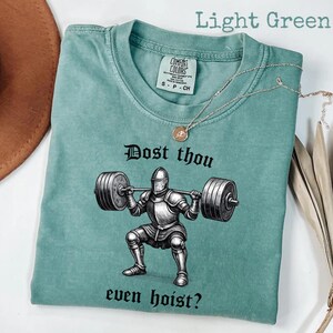 May include: Light green t-shirt with a graphic of a knight lifting weights, and the text "Dost thou even hoist?" A silver necklace with a pendant is draped across the shirt. The shirt has a Comfort Colors tag.