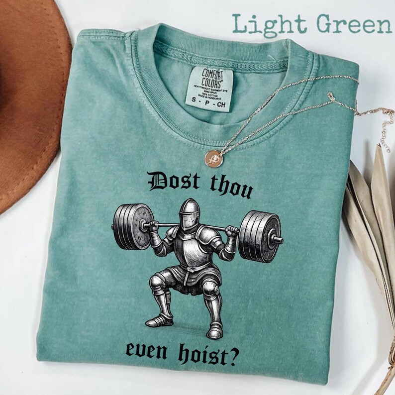 May include: Light green t-shirt with a graphic of a knight lifting weights, and the text "Dost thou even hoist?" A silver necklace with a pendant is draped across the shirt. The shirt has a Comfort Colors tag.