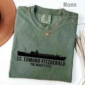 Peut inclure: T-shirt vert mousse avec un motif noir du navire SS Edmund Fitzgerald et le texte "SS. EDMUND FITZGERALD THE MIGHTY FITZ". Le t-shirt a un col rond et des manches courtes. Un collier en or avec un pendentif est drapé sur le t-shirt.
