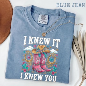 May include: A blue jean colored t-shirt with a graphic print featuring a cowboy hat, pink cowboy boots, horseshoes, and cacti. The text reads I KNEW IT I KNEW YOU. The shirt has a Comfort Colors label at the neck and a gold chain necklace rests on it.
