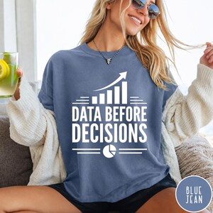 K&ouml;nnte beinhalten: Ein blaues T-Shirt mit Rundhalsausschnitt und wei&szlig;em Aufdruck "DATA BEFORE DECISIONS", erg&auml;nzt durch eine Grafik eines steigenden Balkendiagramms und ein Tortendiagramm-Symbol. Das T-Shirt wird unter einer cremefarbenen Strickjacke getragen.