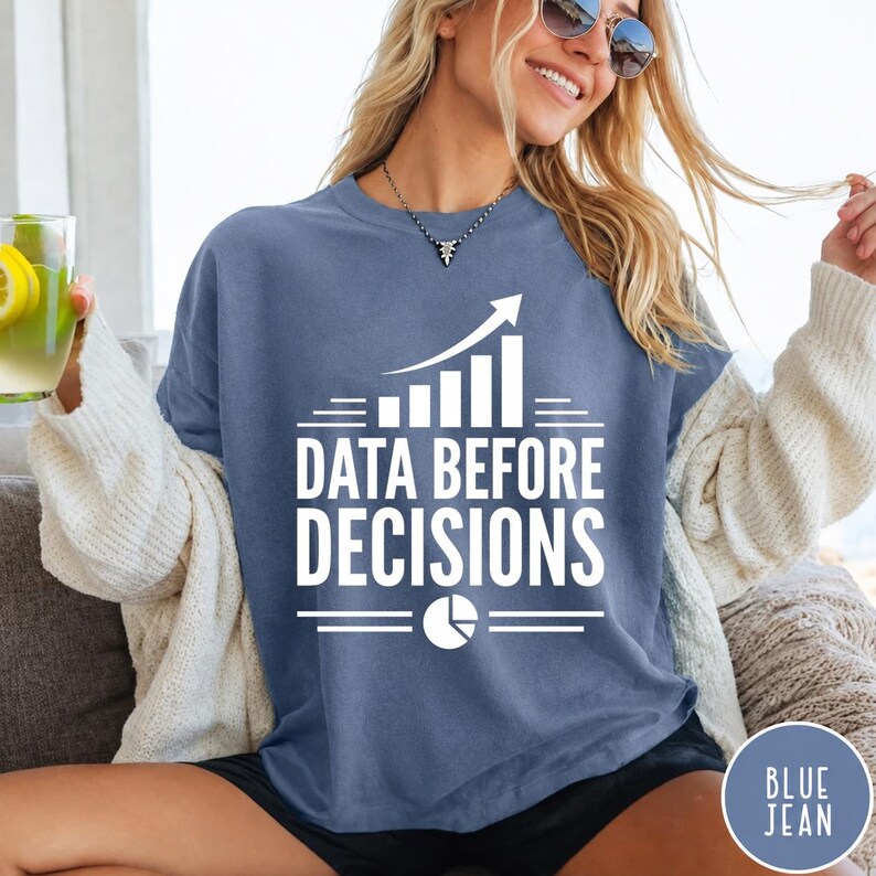 K&ouml;nnte beinhalten: Ein blaues T-Shirt mit Rundhalsausschnitt und wei&szlig;em Aufdruck "DATA BEFORE DECISIONS", erg&auml;nzt durch eine Grafik eines steigenden Balkendiagramms und ein Tortendiagramm-Symbol. Das T-Shirt wird unter einer cremefarbenen Strickjacke getragen.