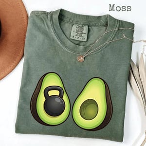Puede incluir: Una camiseta verde musgo con un gráfico de dos mitades de aguacate, una con una pesa rusa negra en su interior. La camiseta tiene una etiqueta Comfort Colors y un collar plateado con un colgante. La palabra "Moss" está impresa en la parte superior derecha.