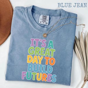 Puede incluir: Una camiseta azul vaquera Comfort Colors con el texto "IT'S A GREAT DAY TO BUILD FUTURES" en letras coloridas. Un collar de oro con un colgante est&aacute; drapeado sobre la camiseta. La camiseta est&aacute; doblada y expuesta sobre una superficie blanca.