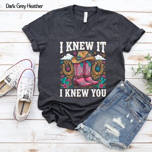 May include: A dark grey heather V-neck t-shirt featuring a graphic print of a cowboy hat, pink western boots, horseshoes, and flowers. The text "I KNEW IT" and "I KNEW YOU" is printed in white above and below the colorful western-themed illustration.