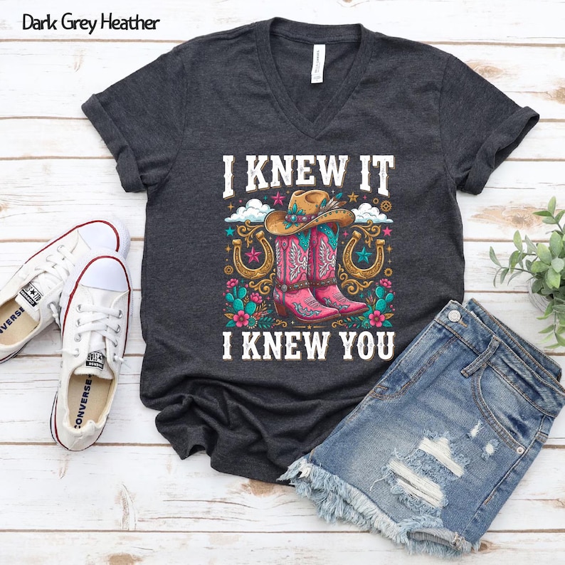 May include: A dark grey heather V-neck t-shirt featuring a graphic print of a cowboy hat, pink western boots, horseshoes, and flowers. The text "I KNEW IT" and "I KNEW YOU" is printed in white above and below the colorful western-themed illustration.