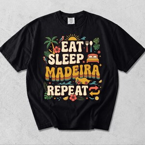 Puede incluir: Una camiseta negra de manga corta con un estampado gráfico colorido que incluye el texto EAT SLEEP MADEIRA REPEAT. El diseño presenta ilustraciones de una palmera, una cama, un tenedor, una cuchara, flores tropicales, un mapa y soles.