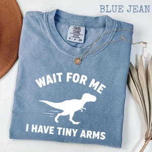 Puede incluir: Camiseta azul vaquero Comfort Colors con el texto "Wait for me, I have tiny arms" y un gráfico de dinosaurio blanco. Un collar de oro con un colgante está sobre la camiseta.
