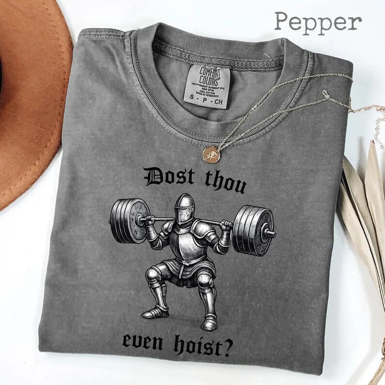 May include: Pepper-colored t-shirt with a graphic of a knight lifting weights, and the text "Dost thou even hoist?" A silver necklace with a pendant is draped across the shirt.