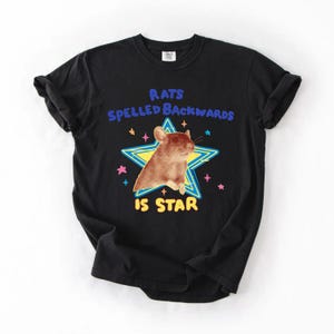 May include: Black t-shirt featuring a brown rat inside a yellow and blue star. The text above the rat reads "RATS SPELLED BACKWARDS" and below the rat, the text reads "IS STAR". Pink and blue stars surround the main graphic.