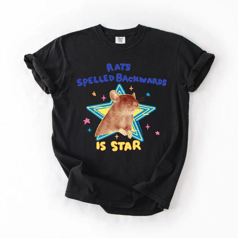 May include: Black t-shirt featuring a brown rat inside a yellow and blue star. The text above the rat reads "RATS SPELLED BACKWARDS" and below the rat, the text reads "IS STAR". Pink and blue stars surround the main graphic.