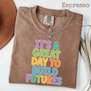 Puede incluir: Camiseta color espresso con el texto "IT'S A GREAT DAY TO BUILD FUTURES" en letras de colores. La camiseta tiene cuello redondo y mangas cortas. Un collar plateado con un colgante est&aacute; sobre la camiseta.