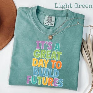 Puede incluir: Camiseta verde claro con el texto "IT'S A GREAT DAY TO BUILD FUTURES" en letras coloridas. La camiseta tiene cuello redondo y mangas cortas. Un collar plateado con un colgante est&aacute; drapeado sobre la camiseta.