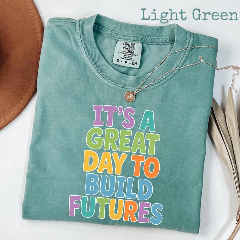 Puede incluir: Camiseta verde claro con el texto "IT'S A GREAT DAY TO BUILD FUTURES" en letras coloridas. La camiseta tiene cuello redondo y mangas cortas. Un collar plateado con un colgante est&aacute; drapeado sobre la camiseta.