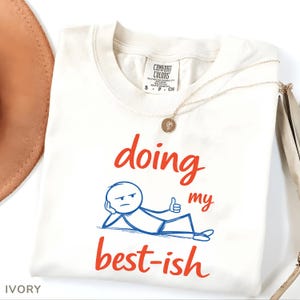 May include: Ivory-colored Comfort Colors t-shirt with the phrase "doing my best-ish" in red and blue. The graphic features a stick figure with a thumbs up. A gold necklace with a pendant is draped across the shirt.