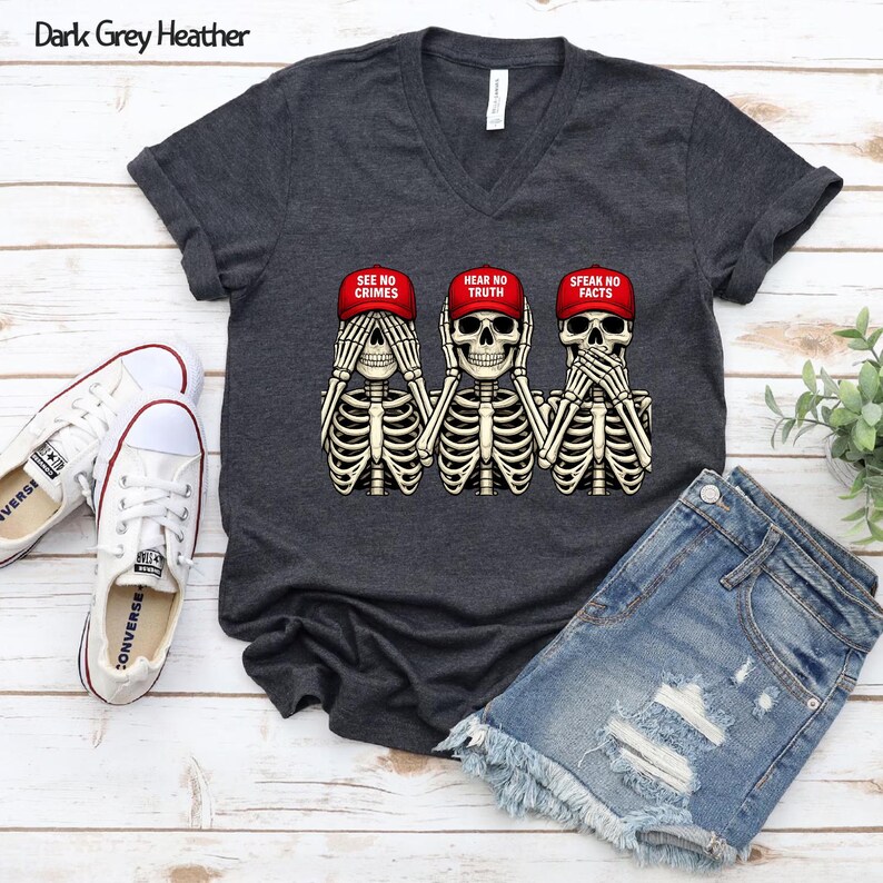 May include: Dark grey heather V-neck t-shirt featuring a graphic of three skeletons wearing red caps, each covering their eyes, ears, and mouth. The caps read "SEE NO CRIMES", "HEAR NO TRUTH", and "SPEAK NO FACTS".